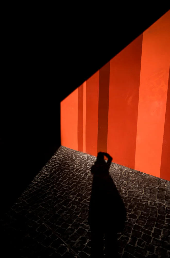 A figure beside a vivid orange wall in deep shadow. Photograph by Adelina Persa.